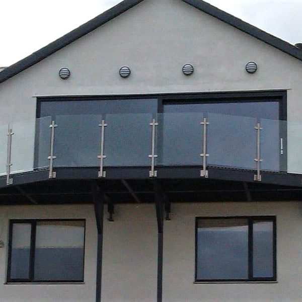 A modern walk-on structural steel balcony with glass panels and stainless steel posts, attached to a two-story house, enhancing the building's exterior with a sleek and contemporary design