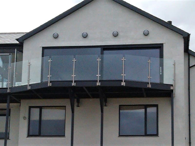 A modern walk-on structural steel balcony with glass panels and stainless steel posts, attached to a two-story house, enhancing the building's exterior with a sleek and contemporary design
