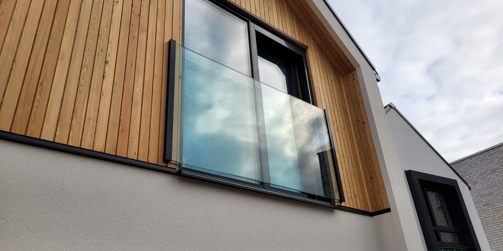 Modern house with a frameless Juliet balcony featuring clear glass panels, adding a sleek and contemporary touch to the wooden facade, enhancing light and ventilation for the upper floor room.