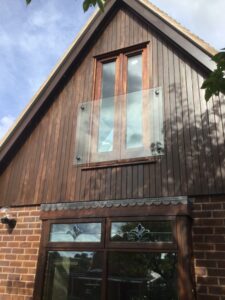 Frameless glass Juliet balcony installed on a modern home, featuring clear toughened safety glass secured with stainless steel adaptors, blending seamlessly with the home's architectural style.